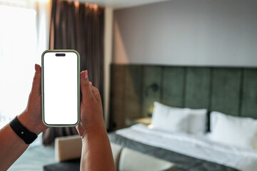Person holding smartphone with blank white screen in hotel room with bed and curtains. Modern...