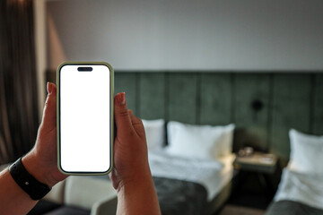 Person holding smartphone with blank white screen in hotel room with bed and curtains. Modern...