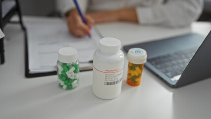 Woman writing on paper at clinic desk with bottles of pills and open laptop in the foreground,...