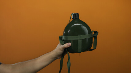 Hand of a man holding a military canteen against an orange background, highlighting the green...