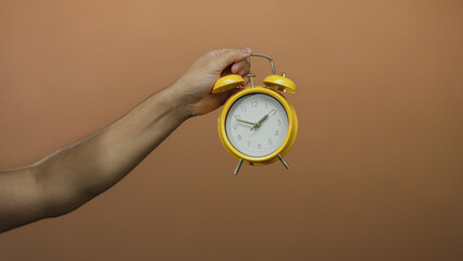 Caucasian man's hand holding yellow alarm clock against brown wall, pointing, isolated, showing...