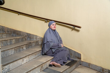 A Muslim woman in a modest gray hijab sits on the steps of a semicircular staircase