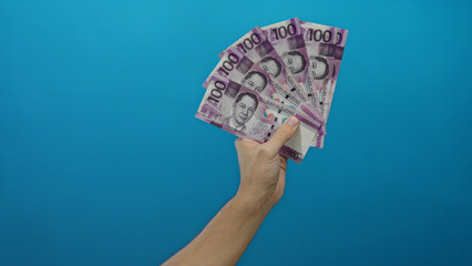 Caucasian man's hand holding philippine pesos against a blue wall, displaying wealth and currency...