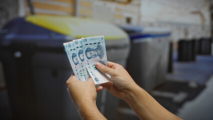 Man holding chinese yuan banknotes beside a recycling bin on a city street, suggesting a blend of...