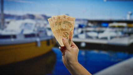 Hand holding brazilian banknotes in an outdoor seaside setting with boats visible in the...