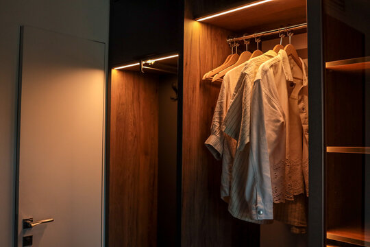 Modern wooden wardrobe with LED lighting and several light shirts on hangers. Minimalist interior design with open closet and contemporary door