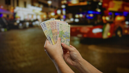 Man holding romanian leu outdoors at a bus stop, showing colorful banknotes against a blurred...