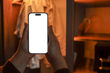 Person holding smartphone with blank white screen in front of wardrobe with clothes on hangers....