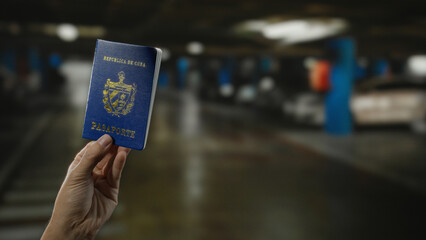 Hand holding cuban passport indoors in a dimly lit parking garage with blurred car in the...