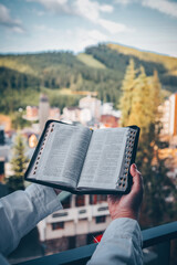 Person holding open Bible on balcony with mountain view and city buildings in background. Concept...