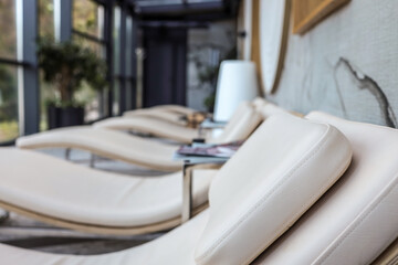 Row of modern spa recliners with soft beige cushions, minimalist side tables and bright natural light through glass walls. Cozy relaxing area in wellness center interior