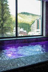Indoor spa pool with glowing purple lights and stone edge, large window with mountain and forest...
