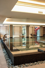 Indoor spa pool with clean water, stone edge, pebble border, lounge chairs, and modern ceiling lighting. Elegant wellness interior with relaxing atmosphere