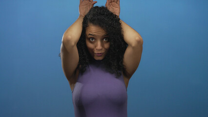 Young woman with bare shoulder makes bunny ears with hand to head in isolated blue studio;...