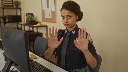 Young woman police officer holds palms forward in a calm gesture at station desk; calm professionalism.