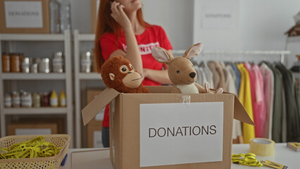 Woman organizing donations in charity room with stuffed animals in box, wearing volunteer shirt, talking on phone, surrounded by clothing and supplies in indoor setting.