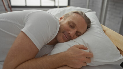 Middle-aged man sleeping peacefully in bedroom, lying comfortably on bed with a white pillow, showcasing a relaxed and serene expression in a calm indoor setting.
