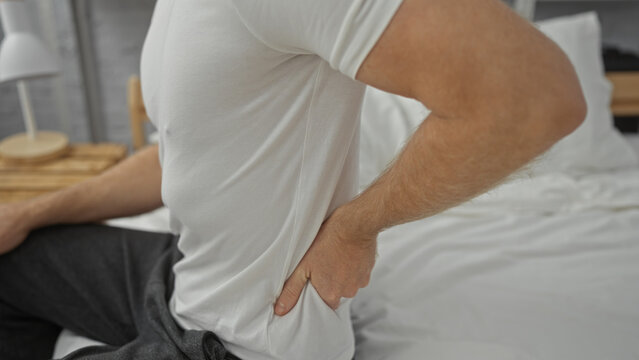 Man holding lower back in discomfort on a bed, wearing casual clothing, positioned indoors, suggesting back pain relief or morning stiffness in a home bedroom setting.