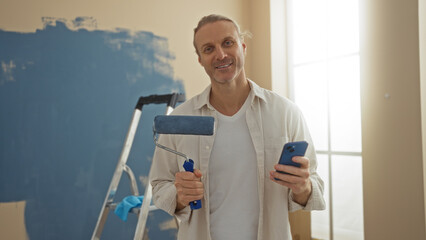 Man painting wall in new home interior holding paint roller and smartphone with ladder in background, showing new apartment renovation in bright living room.