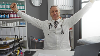 Blonde middle-aged man celebrating success in clinic workspace with arms raised surrounded by...