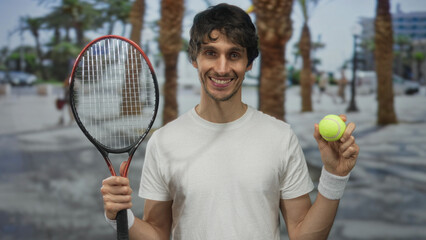 Smiling young man holds tennis racket and ball on sunlit street lined with palm trees wearing a...