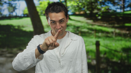 Hispanic man making throat slash gesture exposing neck at a sunlit park with blurred green trees behind him; threat.