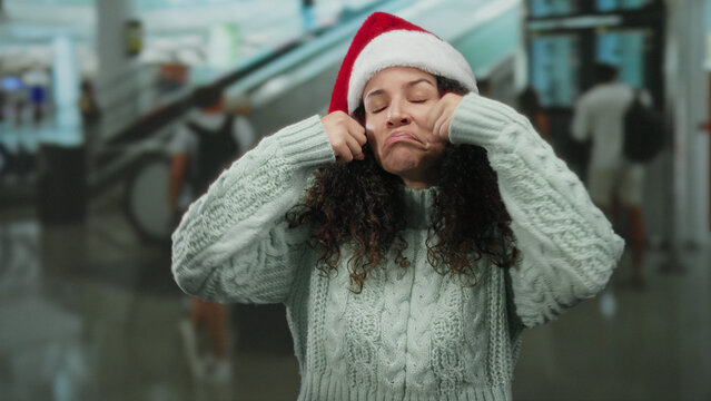 Woman wearing santa hat making crying gesture in airport terminal surrounded by people walking, expressing emotion during holiday travel indoors.