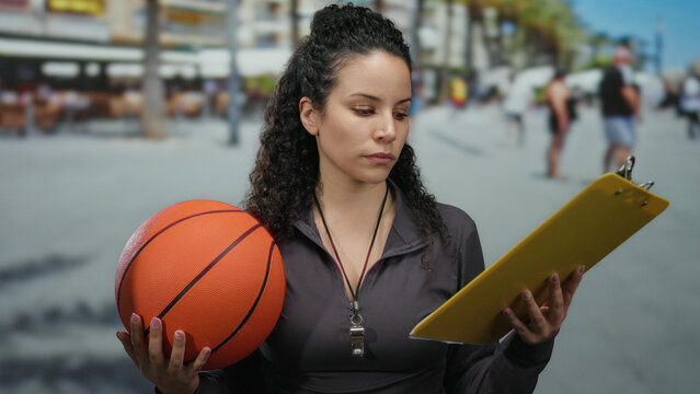Woman holding basketball and clipboard outdoors on a busy street, focused, wearing sports attire, with blurred people in the background, embodying sports leadership.