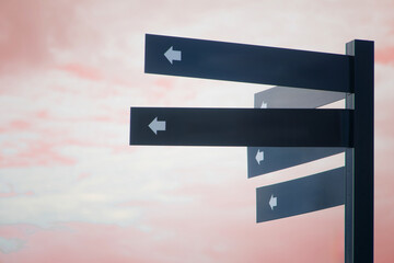 Close-up of a black directional signpost with multiple arrow boards against a red sunset sky,...