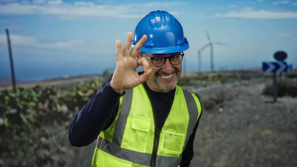 Mature man wearing hardhat and vest gesturing okay in field with windmill energy background showing outdoor job context.