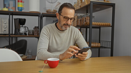 Mature hispanic man in office setting using smartphone with a red cup on the table, surrounded by shelved items.
