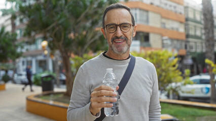 Middle-aged man holding bottle standing outdoors in urban park area with background of city...