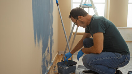 Middle-aged man painting a wall in a new living room, wearing gloves and jeans, using a roller,...
