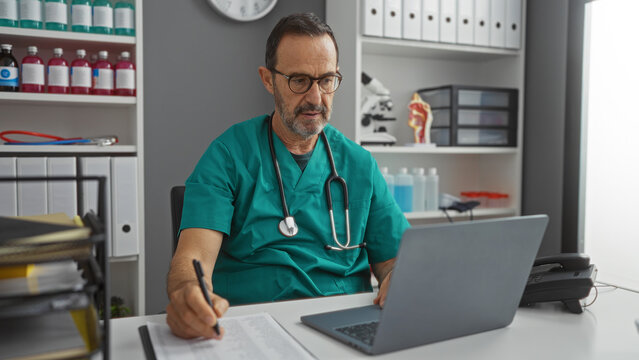 Mature hispanic male doctor working in a hospital clinic room, wearing scrubs and glasses, focused on a laptop while taking notes in an organized indoor environment. - Powered by Adobe