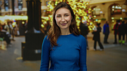 Fototapeta premium Woman smiling on city street during holiday night with bright lights and festive ambiance, young woman in blue sweater enjoying vibrant urban atmosphere outdoors.