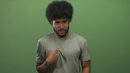 African american man in a gray shirt points to himself against a green wall background, expressing...