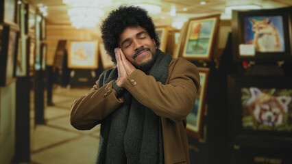 Man with closed eyes gesturing sleep in an art gallery surrounded by paintings and soft lighting,...