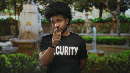 African american man in a security uniform gestures thoughtfully in a green park, with a fountain...