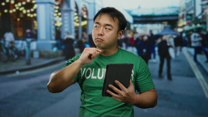 Young man volunteering outdoors in a bustling city street, wearing green shirt, holding tablet,...