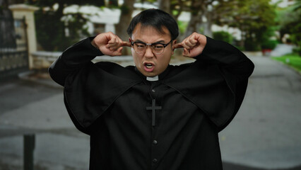 Man wearing glasses and clerical attire stands outdoors on a quiet street, hands covering ears with...