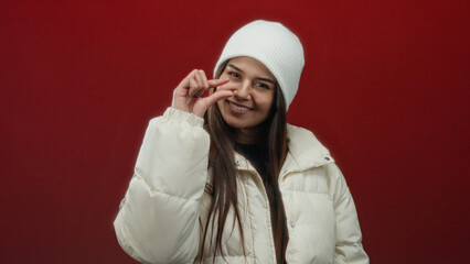 Woman in white coat and beanie gesturing on red background exudes warmth and style while expressing...