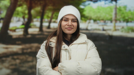 Woman smiling in a winter coat and white beanie against a serene park backdrop with lush green...