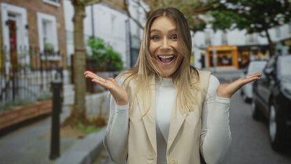 Fototapeta premium Woman smiling with arms raised in surprise on city street background with trees and buildings, wearing white top and beige vest, showcasing joyful expression outdoors.
