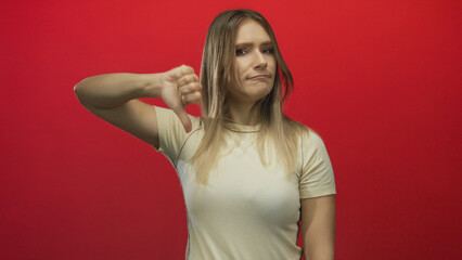 Woman expressing disapproval with a thumbs-down gesture against a bold red background, exhibiting...