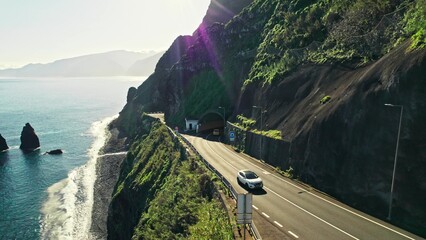 The drone is flying along the mountains on the seashore. A highway with a tunnel on the...