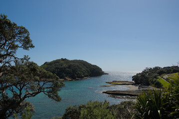 A vast coastal landscape features a long sandy beach, a turquoise ocean with distant islands, and rolling green hills under a bright blue sky with scattered clouds.