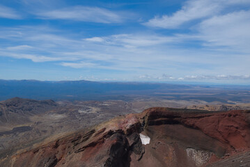 Expansive view over the dramatic, multi-colored volcanic terrain of the Tongariro Crossing, extending to a distant horizon under a wide blue sky.