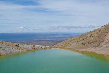 An aerial view of a volcanic landscape with vibrant green and turquoise crater lakes, surrounded by rugged terrain and a trail with hikers, under a clear sky.