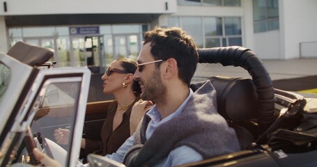 A happy man and a woman are talking animatedly while sitting in a vintage car on a sunny day