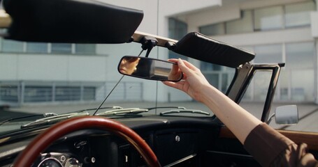 A woman is sitting in the driver's seat of a convertible. She looks in the rearview mirror. A sunny...